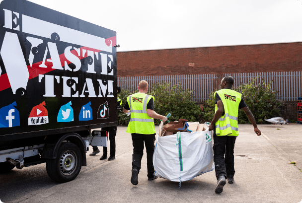 two men with The Waste Team branded high vis jackets on carrying a building waste sack to a waste disposal van