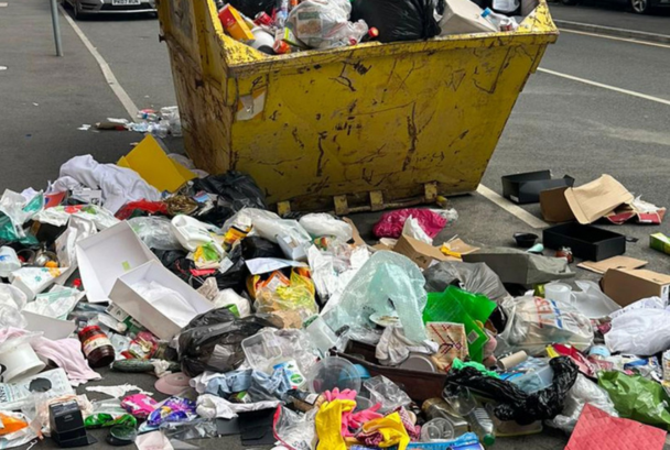 An overloaded yellow skip with fly-tipped rubbish all around it