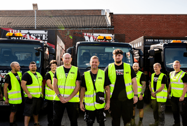The Waste Team workers all lined up wearing branded t-shirts and high vis jackets in front of waste disposal vans