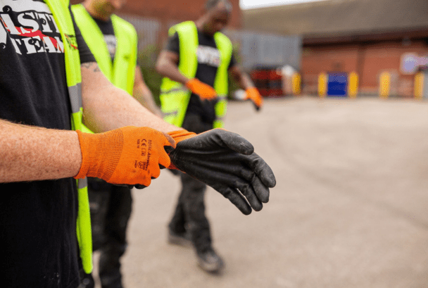 Two men in high-vis jackets and branded The Waste Team t-shirts putting garden waste gloves on