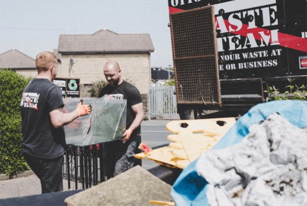 two men in The Waste Team branded t-shirts carrying a box of recyclables go a waste disposal van