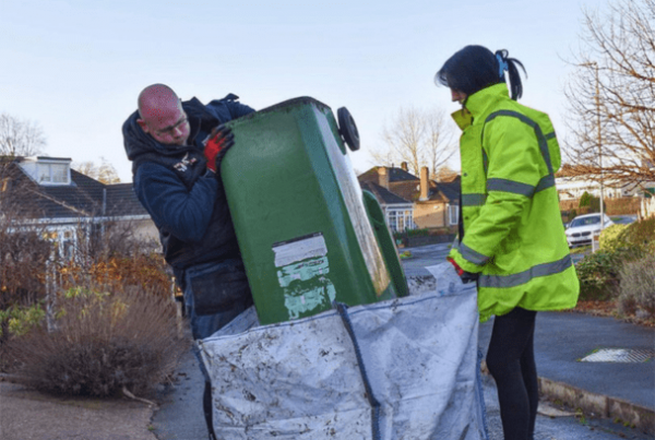 Green recycling bin being emptied by two men into a waste bag