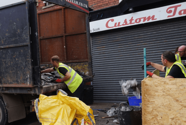 Workers in high vis jackets loading waste disposal van outside shop with shutters down